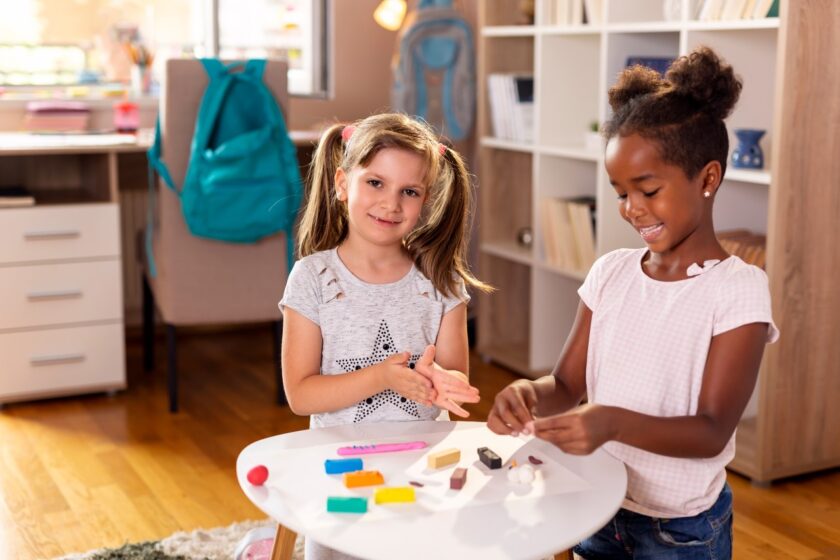 Two young girls stand at a small table in a classroom, smiling and playing with colorful modeling clay. Shelves and a backpack are visible in the background.